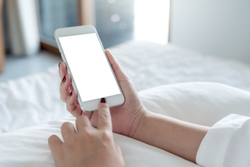 Mockup image of a woman's hand holding and touching white mobile phone with blank desktop screen sitting in the white bed