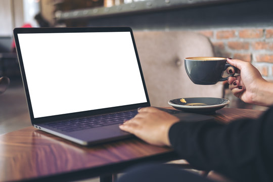Mockup image of a businesswoman using laptop with blank white desktop screen while drinking hot coffee on wooden table in cafe