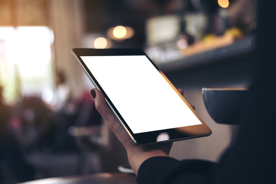 Mockup image of woman's hands holding black tablet pc with blank white desktop screen and coffee cup in cafe background