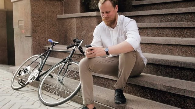Slow motion video of smiling bearded young man sitting on stairs on street and typing message on smartphone