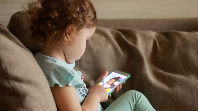 Funny child looks at the phone screen and plays downloaded application on a smart phone close-up. A little cute girl lies in sofa in a living room, looking cartoon and playing the game.