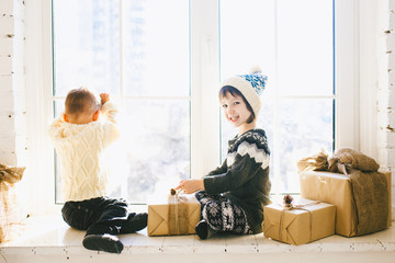 Children brother and sister of preschool age sit by window on a sunny Christmas day and play with gifts boxes wrapped in paper.They are dressed knitted warm woolen clothes and hat.Inside the house