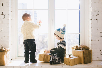 Children brother and sister of preschool age sit by window on a sunny Christmas day and play with...