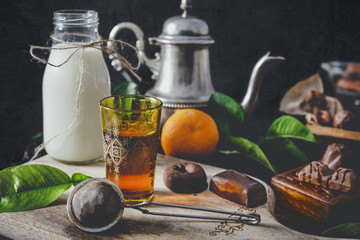 Tea with orange in oriental glass. Milk in glass jar and chocolate sweets. Dark wall at background, copy space.