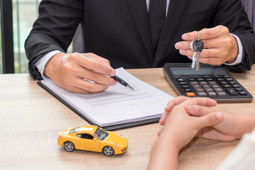 Businessman giving car key and explain about loan agreement with car model on wooden desk.