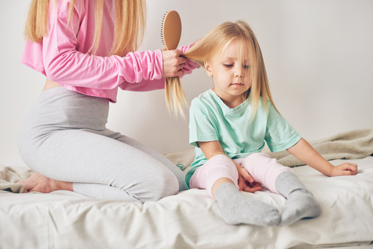 Young mother and daughter doing hair in the morning in bedroom on sunny bright day