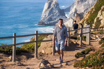 Fototapeta na wymiar Stylish, handsome guy walking along a gravel road against a background of sea rocks (the place where the earth ends and the sea begins, Cabo da Roca (Cape Roca), Sintra, Portugal)