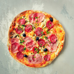 Fresh italian pizza with mushrooms, ham, tomatoes, cheese, olive, pepper on grey concrete background. Copy space. Top view. Square crop