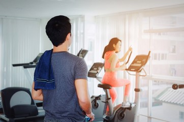 Fototapeta premium muscular young asian man with towel looking to asian young woman slim body exercising on bicycle machine in fitness gym, bodybuilder, healthy lifestyle, fitness, workout and sport training concept