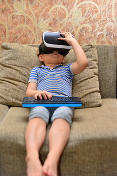 Low angle portrait of little boy wearing VR glasses watching 360 videos and playing augmented reality games at home sitting cross legged on cozy sofa, copy space