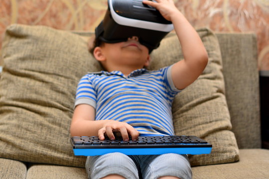 Low angle portrait of little boy wearing VR glasses watching 360 videos and playing augmented reality games at home sitting cross legged on cozy sofa, copy space