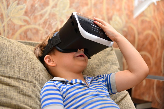 Low angle portrait of little boy wearing VR glasses watching 360 videos and playing augmented reality games at home sitting cross legged on cozy sofa, copy space
