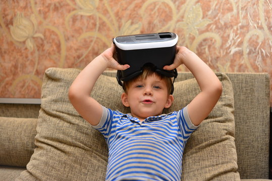 Low angle portrait of little boy wearing VR glasses watching 360 videos and playing augmented reality games at home sitting cross legged on cozy sofa, copy space
