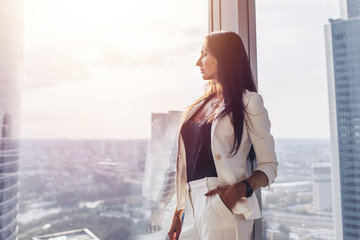 Portrait of elegant business lady wearing white formal suit standing near window looking at cityscape