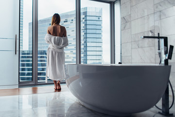 Back view of young woman wearing white bathrobe standing in bathroom looking out the window with bathtub in foreground - Powered by Adobe