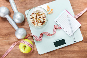 Healthy diet, fitness and weight loss concept. Scales, measuring tape, apple, oatmeal, open notebook and pencil on the table. View from above.
