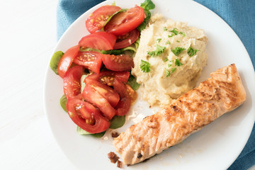 roasted salmon fillet with parsnip puree, tomato salad and parsley garnish, healthy diet meal on a white plate and blue napkin, high angle view from above, copy space