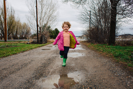 Brother and sister run and splash in puddles in a dirt driveway.