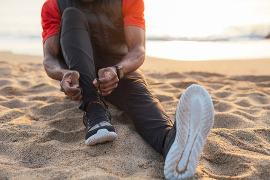 Young Black Athlete Sitting On The Beach At Tying His Shoe At Sunrise.