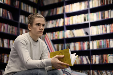 Young Caucasian guy wearing gray sweater spending day at bookstore searching for book on art, holding yellow one in his hands, having uncertain look. People, reading, leisure and knowledge concept
