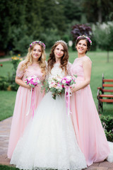 Obraz na płótnie Canvas Stunning young bride and bridesmaids in pink dresses stand with wedding bouquets in the park