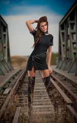 attractive woman with short black dress and long leather boots standing on the rails with bridge in background. Fashion  sexy girl , on the bridge posing in  black long boots on high heels
