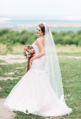 Fototapeta na wymiar Beautiful bride in classy white dress poses with red wedding bouquet on a green lawn