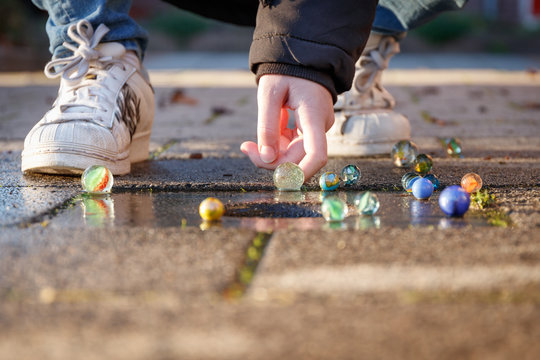 Child is playing with marbles on the sidewalk in the light of the setting sun, it is wearing cool white shoes and a black winter coat.