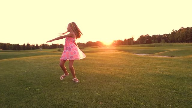 Beautiful child is spinning on lawn at nature. Slow motion cute little girl with long hair is having fun and enjoying nature on green meadow at sunset. Happy active kid.