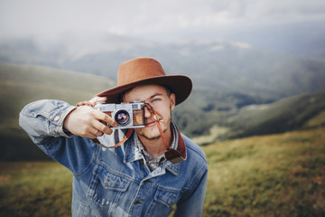 Fototapeta premium stylish man traveler in hat with photo camera smiling on top of mountain. stylish hipster taking photo. space for text. atmospheric moment. wanderlust and travel concept