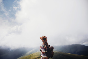 wanderlust and travel concept. girl traveler in hat with backpack looking at clouds in mountains....