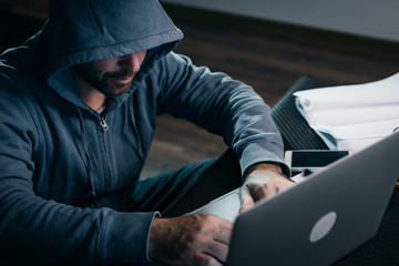 A hacker in a hooded sweatshirt sits behind a laptop