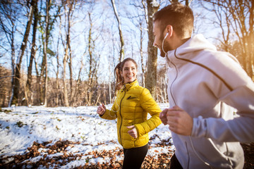 Side view of young pretty charming cheerful fitness shape girl with earphones in winter sportswear jogging in the snowy forest with her boyfriend or trainer.