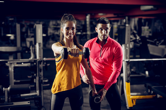 Young healthy motivated happy sporty flexible shape girl doing shoulder exercises with dumbbells in sportswear while handsome helpful personal trainer standing next to her in the gym.