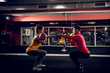 Young healthy focused sporty flexible shape girl with a ponytail doing squat exercises with the kettlebell while handsome helpful personal trainer crouching in front of her.
