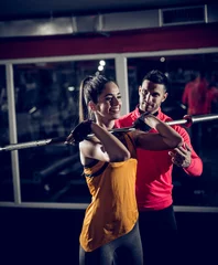 Foto op Canvas Young smiling healthy sporty strong active shape girl holding barbell without weights on the shoulder in front while helpful personal trainer standing next to her. © dusanpetkovic1
