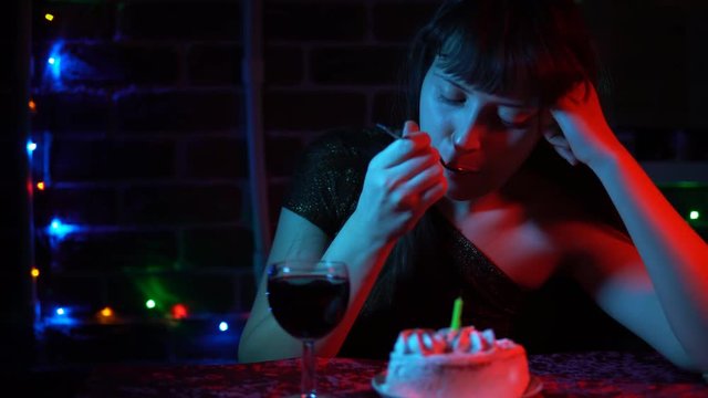 Young attractive woman in a shiny dress drinking wine sitting at a table with a festive cake. Sad brunette celebrates the holiday.