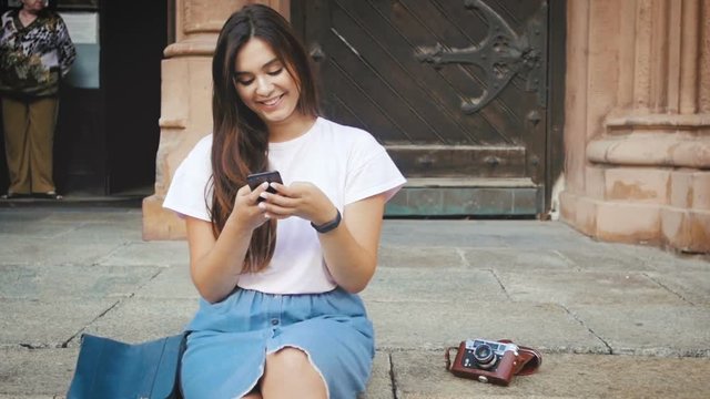 Slow motion footage of smiling young girl sitting on old stone stairs and using mobile phone