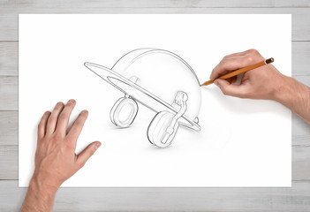 Two male hands draw a pencil sketch of an industrial hard hat with earmuffs on white paper.