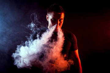 Young stylish male smoker in black T-shirt puffs out of his nose a large cloud of smoke from vape on a black isolated background