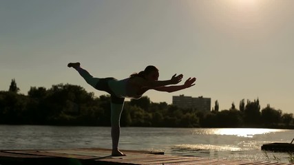 Silhouette of a beautiful healthy girl that does yoga on a wooden bridge near a lake in the nature outside the city. Wide shot.
