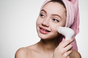 cute smiling girl with a pink towel on her head doing a deeper cleansing of the skin of her face with an electric brush