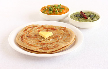 Kerala paratha, a type of Indian bread, with butter topping, and in the background are bowls of peas and carrot curry and pudina chutney.