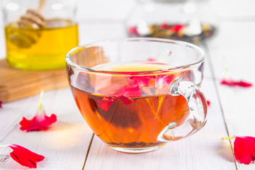 A cup of tea, in the background of a bank of honey and a jar with a black herbal floral tea on a white wooden table.