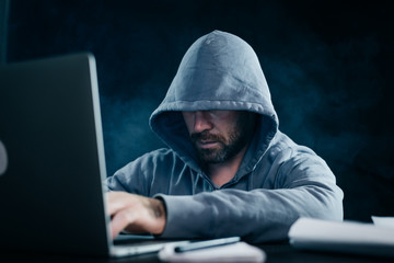 guy hacker with a beard in a sweatshirt with a hood sits behind a laptop
