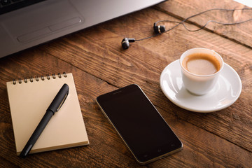  Laptop (notebook) mobile phone (smartphone) and notepad with pen on old wooden table. Work place modern.Cup of coffee
