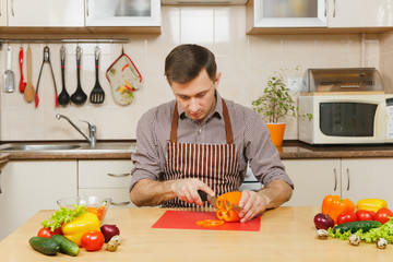 Handsome smiling caucasian young man in an apron, brown shirt sitting at table, cuts vegetable for salad with knife in light kitchen. Dieting concept. Healthy lifestyle. Cooking at home. Prepare food.