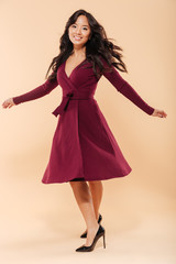 Full size portrait of charming asian female in pretty maroon dress dancing and looking on camera being isolated over beige background