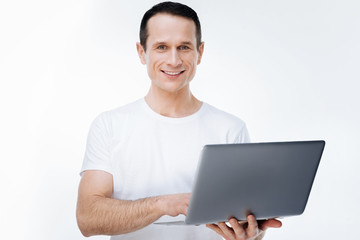 Modern device. Delighted happy nice man smiling and using a laptop while standing against white background