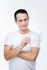 Positive look. Nice positive handsome man standing cross handed and smiling to you while holding his glasses